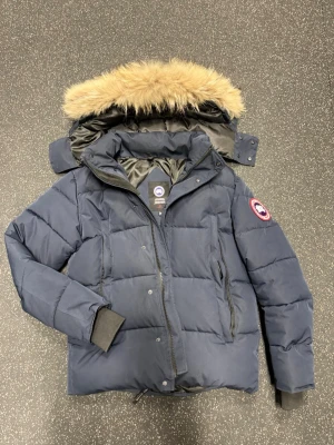 Mörkblå dunjacka från Canada Goose - Säljer en mörkblå dunjacka från Canada Goose med avtagbar huva och snygg pälskant. Jackan har två dragkedjeförsedda fickor, ribbade muddar och klassisk patch på ärmen. Perfekt för kalla vinterdagar och riktigt varm.