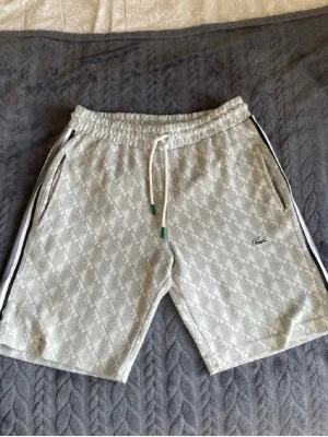 Grå mönstrade shorts från Lacoste - Säljer ett par grå shorts från Lacoste med diskret rutigt mönster och vita sidoremsor. Shortsen har snörning i midjan, fickor på sidorna och en bakficka. Perfekta för sommaren och riktigt sköna att bära.