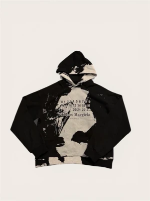 Maison Margiela svart hoodie med tryck - Säljer en svart hoodie från Maison Margiela med unikt vitt konstnärligt tryck och logga på bröstet. Hoodien har huva och är tillverkad i mjuk bomull. Perfekt för dig som gillar streetwear och vill sticka ut med en exklusiv look.
