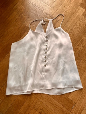 Vit linnetopp med knappar - Säljer en stilren vit linnetopp med tunna axelband och dekorativa knappar framtill. Toppen har v-ringad hals och ärmlös design, perfekt för varma dagar. Materialet känns lätt och svalt, troligen polyester eller liknande.Skriv gärna för mått! 💕