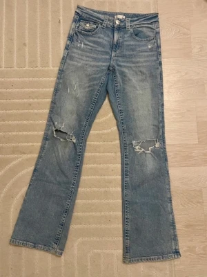Ljusblå slitna bootcut jeans - Säljer ett par ljusblå bootcut jeans med slitningar och hål på knäna. Jeansen har klassisk femficksmodell, knappgylf och dekorativa ficklock bak. Perfekta för en avslappnad och trendig look.