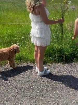 Vit volangklänning med spetsdetaljer - Söt vit kortklänning med flera lager volanger och spetsdetaljer. Klänningen är ärmlös och har en luftig, romantisk känsla. Perfekt för sommarens alla tillfällen och ger en fräsch look.