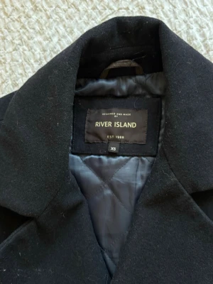 Svart skepparkavaj River Island XS - Klassisk svart skepparkavaj från River Island med dubbelknäppning och breda slag. Jackan har två fickor framtill och är helfodrad med quiltat innerfoder. Perfekt för dig som gillar stilren och tidlös design. Vid snabb affär så kan man få priset lite sänkt 