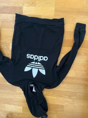 Svart hoodie från Adidas med logga - Säljer en svart hoodie från Adidas med stor vit Trefoil-logga och text på bröstet. Klassisk känguruficka framtill och huva. Tillverkad i mjukt material, perfekt för en avslappnad streetwear-look.