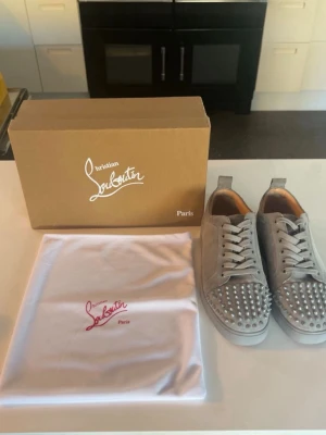Grå Christian Louboutin sneakers med nitar - Snygga grå sneakers från Christian Louboutin i mocka med silvriga nitar på tån och den klassiska röda sulan. Skorna har vita skosnören och en låg profil. Perfekta för dig som vill sticka ut med exklusiv streetstyle.