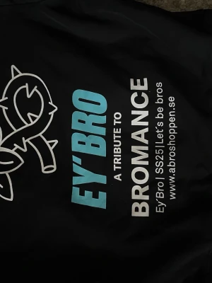 Svart hoodie Ey'Bro med tryck - Svart hoodie från Ey'Bro med stort grafiskt tryck på ryggen i vitt och blått. Texten 'A tribute to bromance' och en stiliserad ros ger en unik vibe. Hoodien har ribbade muddar och klassisk huva. Perfekt för dig som vill sticka ut med streetwear.