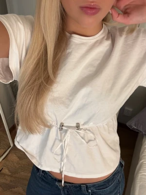 Vit croppad topp med snörning - Säljer en vit croppad topp med korta ärmar och snörning i midjan som ger en snygg passform. Perfekt till jeans eller kjol för en chill och trendig look. Materialet känns mjukt och bekvämt, troligen bomull. Enkel och stilren design. Använd ca 3 ggr så som ny❤️