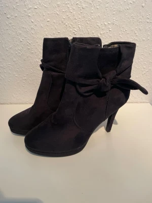 Svarta stövletter med rosett - Snygga svarta stövletter i mockaimitation med hög klack och dekorativ rosett vid ankeln. Dragkedja på insidan för enkel på- och avtagning. Perfekta för dig som vill ha en stilren men ändå unik look.