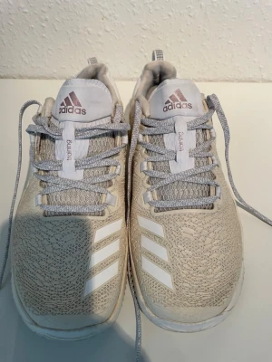 Beige Adidas sneakers med mönster - Snygga beige sneakers från Adidas med vita detaljer och klassiska tre ränder på sidan. Skorna har ett diskret mönster i materialet, gråvita skosnören och Adidas-logga i brons på plösen. Låg modell med bekväm sula och dragflik bak.