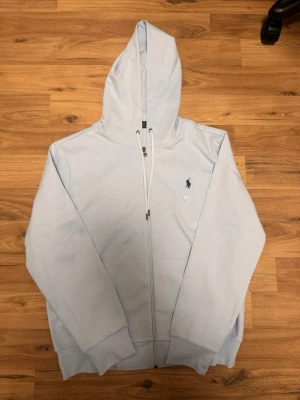 Ljusblå hoodie från Ralph Lauren - Snygg ljusblå hoodie från Ralph Lauren med dragkedja framtill och klassisk Polo-logga broderad på bröstet. Hoodien har justerbar huva med snören och ribbade muddar vid ärmar och nederkant. Perfekt för en avslappnad och stilren look.