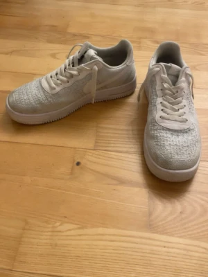 Vita Nike Air Force 1 Flyknit sneakers - Snygga vita Nike Air Force 1 Flyknit sneakers med klassisk låg siluett. Ovandelen är tillverkad i ett stickat material som ger en lätt och luftig känsla. Skorna har vita snören och en vit sula med diskret Nike-logga på sidan.