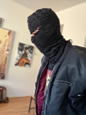 Svart balaclava i spets - Unik svart balaclava i spetsmaterial med hål för ögon och mun. Masken har ett mönstrat tyg som ger en cool och edgy look. Perfekt för dig som vill sticka ut med din stil och gillar accessoarer med attityd.
