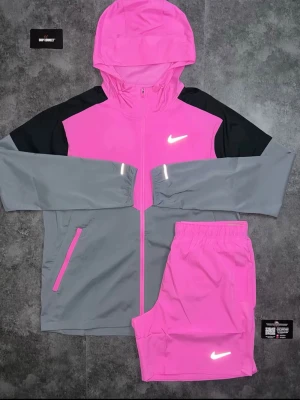 Nike rosa och grå träningsshorts - Snygga träningsshorts från Nike i en stark rosa färg med vit logga. Shortsen har elastisk midja och är tillverkade i ett lätt och funktionellt syntetmaterial som passar perfekt för sport och aktivitet. Matcha med jacka för en komplett look.