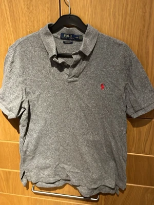 Grå pikétröja från Polo Ralph Lauren - Klassisk grå pikétröja från Polo Ralph Lauren med custom fit. Tröjan har korta ärmar, krage och knappar framtill samt den ikoniska röda loggan broderad på bröstet. Perfekt till jeans eller shorts för en clean och stilren look.