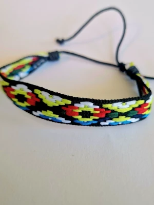 Färgglatt vävt vänskapsarmband - Säljer ett handgjort vänskapsarmband med färgglada geometriska mönster i svart, rött, gult, vitt och blått. Armbandet har justerbar knytning och är gjort av slitstark tråd. Perfekt accessoar för att ge din stil en personlig touch.