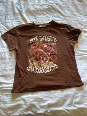 Brun t-shirt med svampmotiv - Cool brun t-shirt med tryck av svampar och blommor samt texten 'Good Thoughts New Possibilities' på framsidan. Skön passform och tillverkad i mjuk bomull, perfekt för en avslappnad stil.