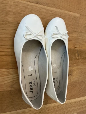Vita ballerinaskor med rosett Jana - Snygga vita ballerinaskor från Jana med rund tå och dekorativ rosett framtill. Skorna har en klassisk och enkel design med mjuk innersula för extra komfort. Perfekta till både jeans och klänning. 