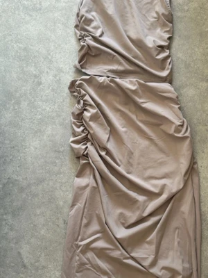 Beige långklänning med rynkade detaljer - Snygg beige set med rynkade partier längs sidorna som ger en cool och modern look. Setet är ärmlös och har en mjuk, stretchig känsla. Perfekt för dig som gillar stilrena och enkla plagg med en twist.
