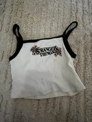 Stranger Things vit linnetopp - Vit linnetopp med svarta kanter och Stranger Things-tryck framtill dekorerat med blommor. Tunn och lätt, perfekt för varma dagar. Axelbandsmodell med croppad passform som ger en trendig look.
