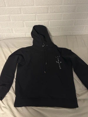 Svart Cactus Jack hoodie - Svart hoodie från Cactus Jack med tryck på bröstet och stor grafisk print på ryggen med texten 'LOOK MOM I CAN FLY'. Hoodien har dragsko i huvan och ribbade muddar. Perfekt för dig som gillar streetwear och Travis Scott-inspirerad stil.