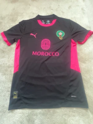 Marocko fotbollströja Puma svart/rosa - Snygg svart fotbollströja från Puma med rosa detaljer och text. Marockos landslagsemblem på bröstet och tryck med 'MOROCCO' framtill. Tillverkad i lätt och ventilerande material, perfekt för träning eller match.