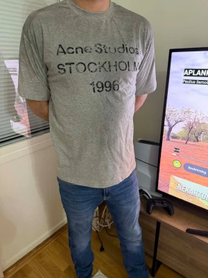 Grå Acne Studios t-shirt med tryck - Snygg grå t-shirt från Acne Studios med svart tryck på bröstet där det står 'Acne Studios STOCKHOLM 1996'. Klassisk passform och rund halsringning. Perfekt för en avslappnad och trendig look.
