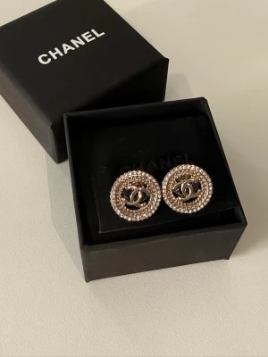 Chan* örhängen med pärlor - Snygga runda örhängen från Chan* med ikoniska loggan i mitten. Örhängena är silverfärgade och har en ram av små pärlor som ger en lyxig känsla. Perfekt för dig som vill ha något exklusivt och trendigt. Levereras i original ask.