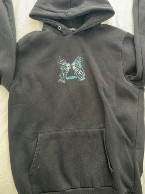 Svart hoodie med fjäril från Aarhon - Cool svart hoodie från Aarhon med stor blå och vit fjäril på både bröstet och ryggen. Hoodien har huva, ribbade muddar och en praktisk magficka. Perfekt för dig som gillar unika prints och streetwear-vibe. Hör av dig vid minsta lilla fråga och priset är absolut inte hugget i sten😄