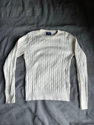 Vit kabelstickad tröja från GANT - Snygg vit kabelstickad tröja från GANT med rund hals och broderad logotyp på bröstet. Tröjan har långa ärmar och ribbade muddar. Perfekt för dig som gillar klassisk och stilren design. Materialet är mjukt och bekvämt.