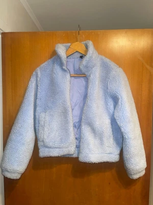 Ljusblå teddyjacka med krage - Mysig ljusblå teddyjacka med hög krage och långa ärmar. Jackan är tillverkad i fluffigt teddy-material och har en enkel, rak passform. Perfekt för att hålla sig varm under kyliga dagar. Jackan saknar synliga fickor och knäppning.