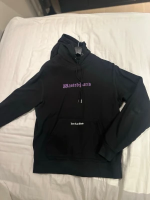 Svart hoodie från Wasted Paris - Svart hoodie från Wasted Paris med lila tryck på bröstet och vit text på fickan. Klassisk huva med snören och stor magficka framtill. Materialet är mjukt och bekvämt, perfekt för en avslappnad streetwear-look.