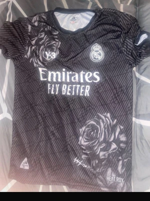 Real Madrid x Y-3 svart matchtröja - Snygg svart Real Madrid matchtröja i samarbete med Y-3 och Adidas. Tröjan har vita rosdetaljer, klubbmärke, sponsortryck och HEAT.RDY-teknologi. På ryggen står det 'MBAPPÉ' och nummer 9 i vitt. Perfekt för dig som älskar fotboll och streetwear.