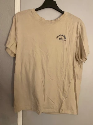 Beige t-shirt med tryck Vancouver - Säljer en beige t-shirt med ett litet tryck på bröstet där det står 'Vancouver 1971'. T-shirten har rund hals och korta ärmar, perfekt för en avslappnad och enkel stil. Materialet känns mjukt och bekvämt, troligtvis bomull.