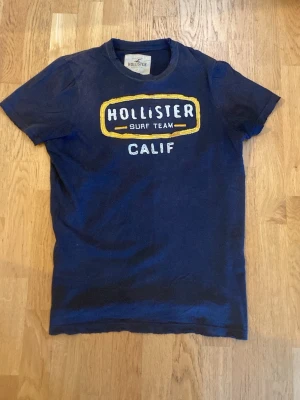 Blå Hollister t-shirt med tryck - Mörkblå t-shirt från Hollister med stort broderat tryck på bröstet där det står 'Hollister Surf Team Calif' i vitt och gult. Klassisk rund hals och korta ärmar. 