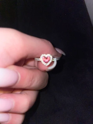 Pandora silverring med rosa hjärta - Säljer en snygg silverring från Pandora med ett hjärtformat motiv i mitten. Hjärtat har en rosa sten och är omgiven av små gnistrande stenar. Ringen har även stenar längs bandet för extra bling. Perfekt för dig som gillar söta och romantiska detaljer. Strl 52