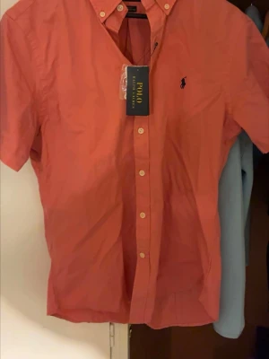 Rosa kortärmad skjorta Polo Ralph Lauren - Snygg rosa skjorta från Polo Ralph Lauren med korta ärmar och klassisk button-down krage. Slim fit-modell i lätt bomullstyg, perfekt för en fräsch och stilren look. Ikoniska loggan broderad på bröstet och knäppning framtill. Bilderna är tagna i ett annorlunda ljus och ser därför mer rött ut, kolla bild TVÅ för rätta färg!