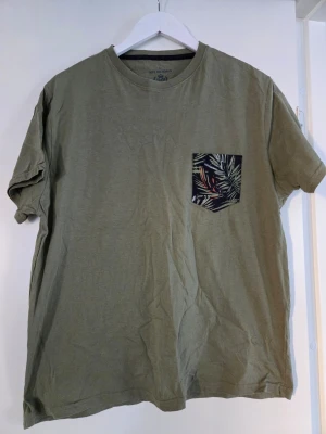 Olivgrön t-shirt med mönstrad ficka - Cool olivgrön t-shirt från Mel Richman i bomull, med en unik bröstficka som har ett tropiskt mönster i flera färger. Perfekt för dig som vill ha en enkel men ändå lite extra detalj på din outfit.