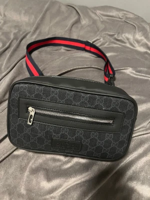 Svart axelväska från GG - Säljer en svart axelväska med klassiskt GG-mönster och läderdetaljer. Väskan har en silverfärgad dragkedja framtill och en bred axelrem i rött och blått. Perfekt för dig som vill ha en stilren och trendig väska.