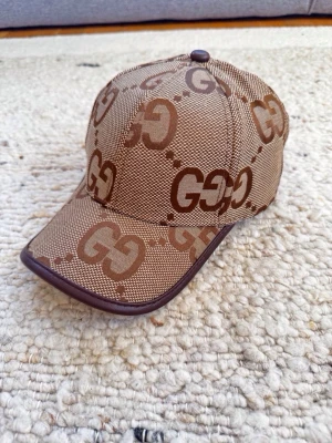 Beige GG Jumbo keps - Snygg brun keps med det klassiska GG-mönstret i olika nyanser av brunt. Kepsen har böjd skärm med mörkbrun kant och justerbar rem bak. Tillverkad i slitstarkt tyg med exklusiv känsla, perfekt för dig som gillar streetwear och designerplagg. Äkta läder på kanterna, made in Italy.