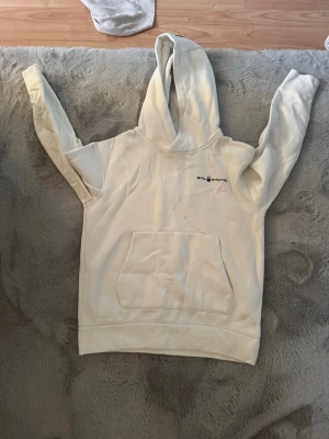 Vit hoodie från Sail Racing - Säljer en stilren vit hoodie från Sail Racing med svart logga på bröstet. Hoodien har en stor känguruficka framtill och klassisk huva. Perfekt för dig som gillar enkel och sportig stil. Materialet är mjukt och skönt, troligtvis bomullsblandning.