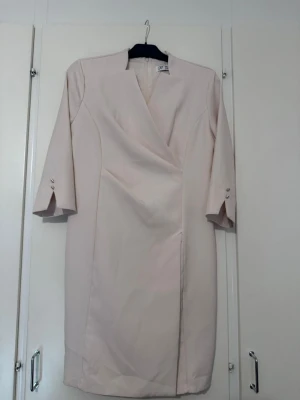 Beige omlottklänning med trekvartsärm - Stilren beige omlottklänning med trekvartsärm och dekorativa knappar vid ärmslut. Klänningen har en rak passform och v-ringad halsringning. Perfekt för dig som gillar en klassisk och elegant look.