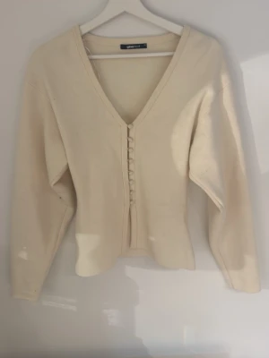 Krämvit kofta med knappar från Gina Tricot - Snygg krämvit kofta från Gina Tricot med djup V-ringning och dekorativa knappar framtill. Koftan har lång ärm och är figurnära i modellen. Perfekt att styla med jeans eller kjol för en clean look.