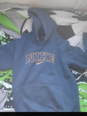 Blå hoodie från Nike med tryck - Blå hoodie från Nike med stort guldfärgat NIKE-tryck på bröstet och klassisk huva med dragsko. Mjuk insida och känguruficka framtill. Perfekt för en avslappnad och sportig stil.