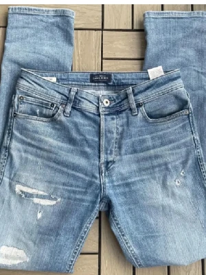 Ljusblå slitna jeans från Jack & Jones - Ljusblå jeans från Jack & Jones med raka ben och slitna detaljer på framsidan. Klassisk femficksmodell med knappgylf och bälteshällor. Jeansen har en trendig tvätt och är tillverkade i denim.