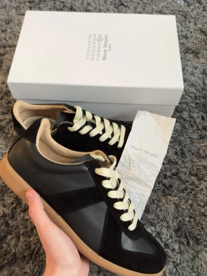Maison Margiela svarta sneakers - Maison Margiela sneakers i svart läder och mocka med beige detaljer och krämvita snören. Klassisk låg modell med gummisula och diskret branding. Perfekt för dig som vill ha en stilren och trendig sneaker.
