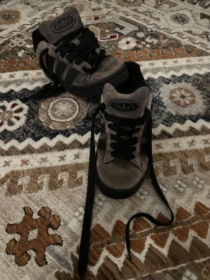 Grå mocka sneakers från Adidas - Snygga sneakers från Adidas campus  i grå mocka med svarta detaljer och klassiska ränder på sidorna. Skorna har svart snörning och Adidas-logga på plösen. Yttersulan är svart och har ett greppvänligt mönster. Sko snöre på höger sida av skon har tyvärr gått sönder lite men sitter fortfarande fast bara att det har gått ur sina flätor, storlek kan också vara 40-41