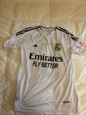 Real Madrid Bellingham matchtröja - Vit Real Madrid matchtröja från Adidas med svarta detaljer och klubbmärke på bröstet. Tryck med 'Emirates Fly Better' på framsidan och 'Bellingham 5' på ryggen. La Liga-märke på ärmen och klassiska Adidas-ränder på axlarna. Perfekt för fotbollsfans.