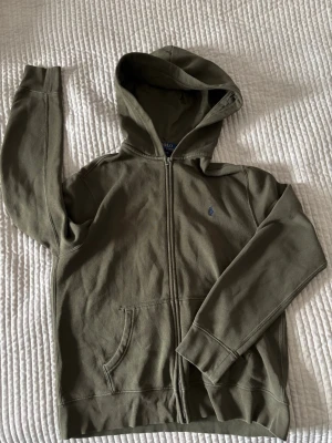 Olivgrön zip hoodie från Polo Ralph Lauren - Säljer en olivgrön hoodie från Polo Ralph Lauren med dragkedja och huva. Tröjan har två stora fickor framtill och ett diskret blått Polo-logo broderat på bröstet. Materialet är mjukt och bekvämt, äkta och passar ca 150cm, storleken på lappen är L(14-16)