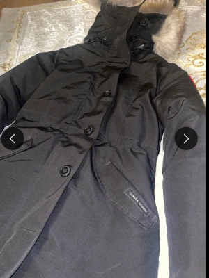 Svart parkas från Canada Goose - Säljer en svart parkas från Canada Goose med huva och snygg pälsdetalj. Jackan har stora knappar framtill, fickor med logotyp och en längre passform som håller dig varm. Perfekt för kalla dagar och har en stilren design som passar till många outfits.
