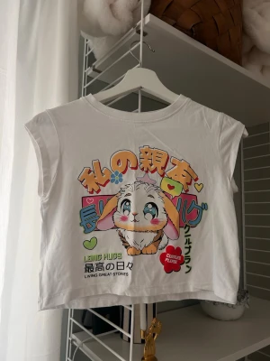 Vit croppad topp med kaninmotiv - Söt vit croppad topp med färgglatt anime-inspirerat kanintryck och japansk text framtill. Toppen är ärmlös och har en avslappnad passform, perfekt för en lekfull och trendig stil.